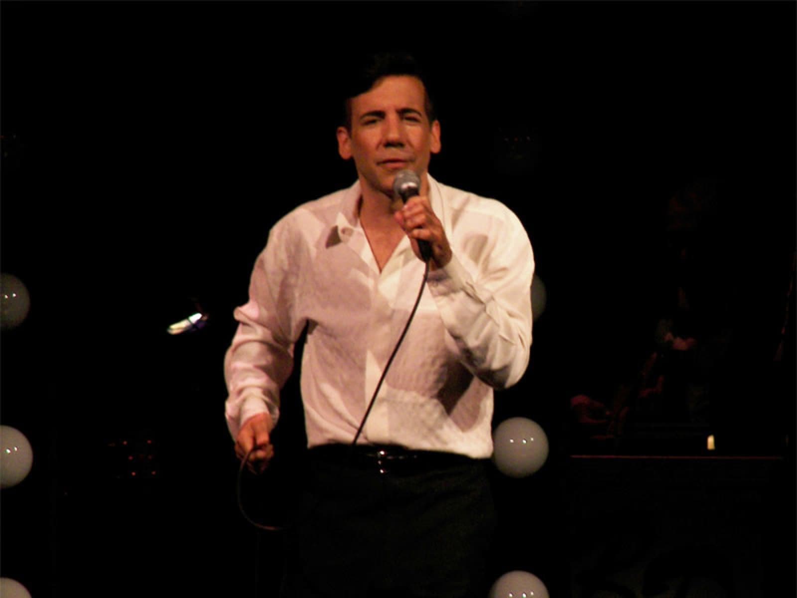 Chaz Esposito in a white shirt singing into a handheld microphone under stage light