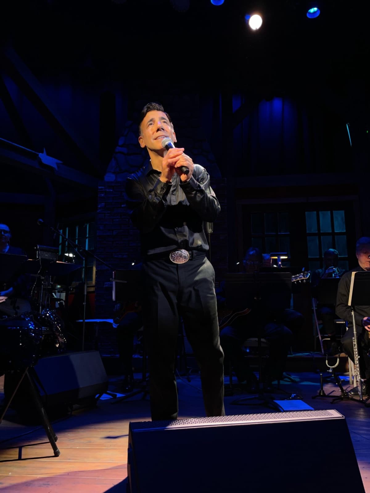 Chaz Esposito in black stage wear singing with the show orchestra behind him in a stone-walled concert hall