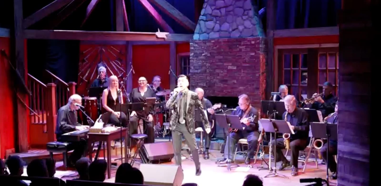 Wide view of Chaz Esposito and the full show band performing on a theatre stage with a stone fireplace backdrop