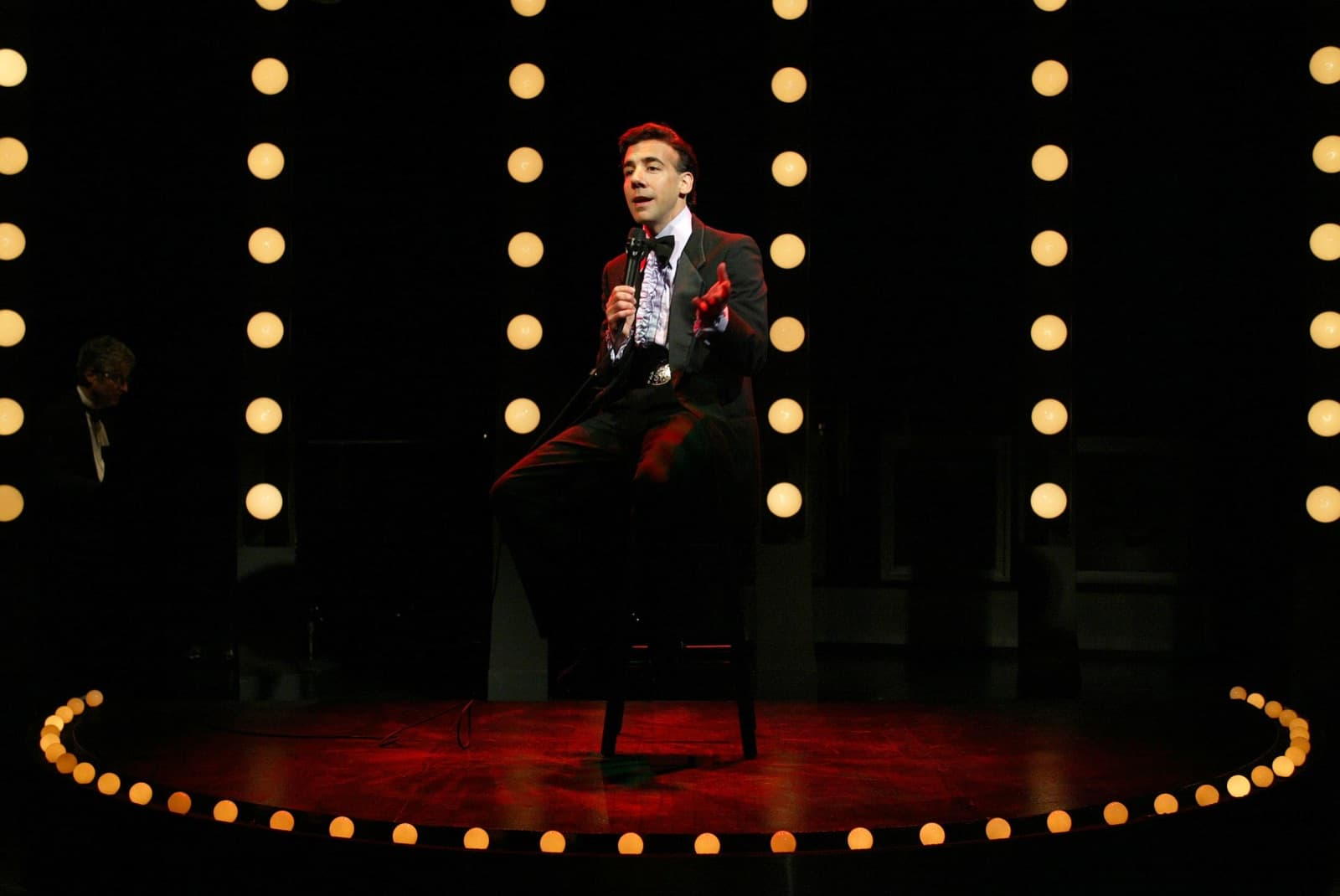 Chaz Esposito as Bobby Darin, seated on a stool in a spotlight against a wall of marquee bulbs
