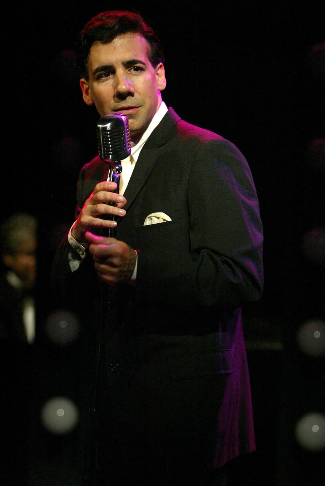 Chaz Esposito holding a vintage chrome microphone stand, pocket square in his dark suit